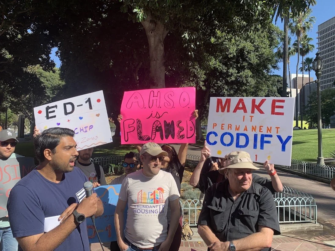 Protesters holding signs, with one speaker speaking into a microphone, in a park with trees and tall buildings in the background.