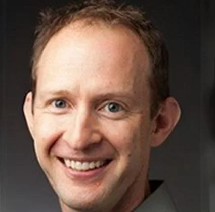 Close-up of a smiling man with light skin, short brown hair, and blue eyes.