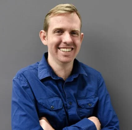 A man with blond hair wearing a blue button-up shirt, smiling and standing with crossed arms against a gray background.
