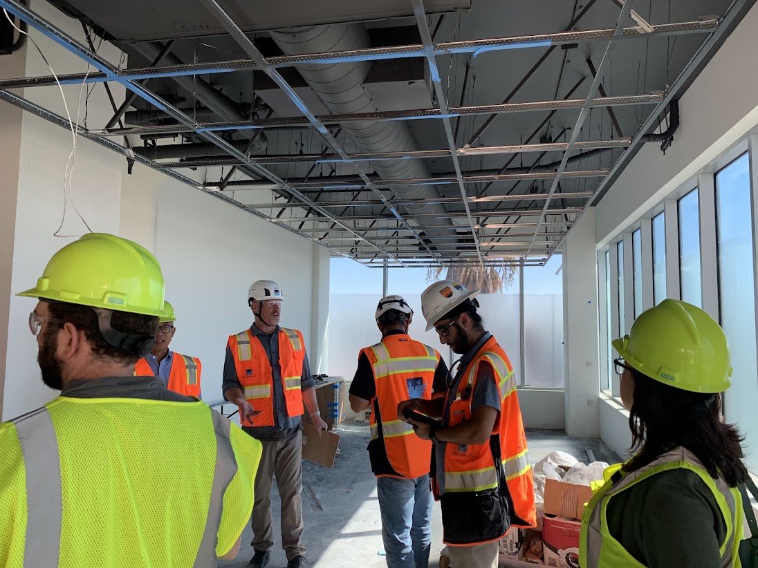 Construction workers and a supervisor in a partially finished building with exposed ceiling framework, large windows, and construction materials.
