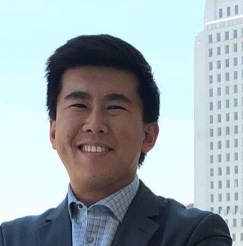 A smiling man in a suit standing outdoors with a tall building in the background.
