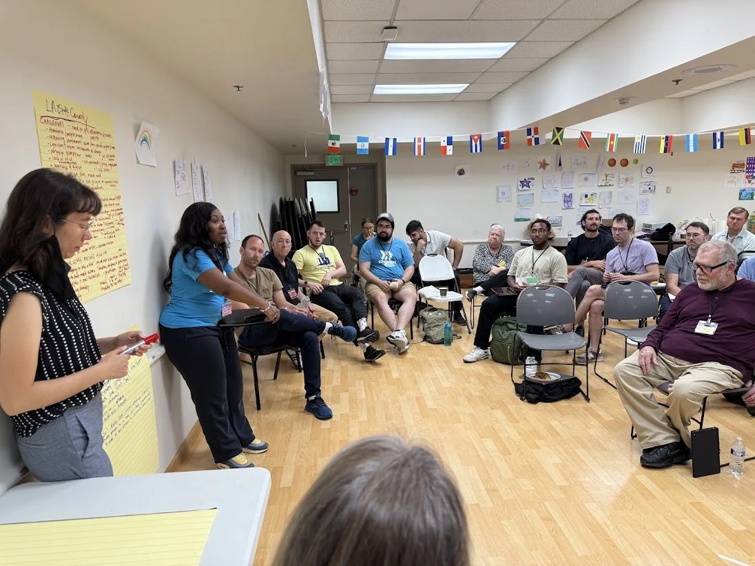 A diverse group of people seated in a semi-circle in a room, participating in a discussion or workshop. Two women are standing at the front, one speaking and the other reading from a notepad. The room has colorful flags hanging from the ceiling, children's drawings on the wall, and large yellow posters with notes.