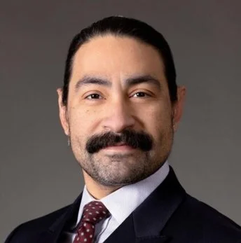 Professional man with dark hair and a mustache, wearing a suit and tie, looking at the camera.