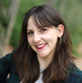 A woman with brown hair, wearing a dark green jacket and hoop earrings, smiling outdoors against a green, blurred background.