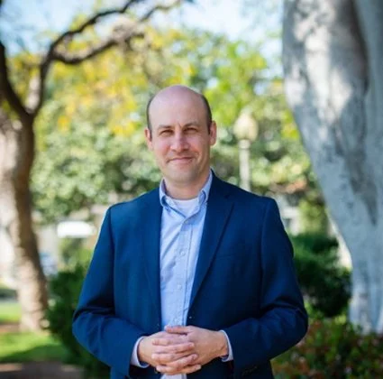 Man in a blue suit and light blue shirt standing outdoors with trees in the background.