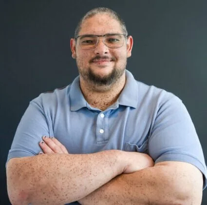 A man with glasses and a beard, wearing a light blue polo shirt, standing in front of a dark background with arms crossed.