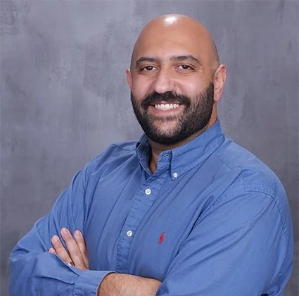 Headshot of a smiling man with a beard, wearing a blue button-up shirt, standing against a gray background.