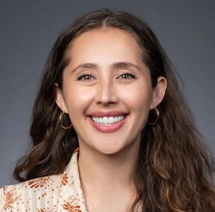Close-up of a smiling woman with long, wavy brown hair, wearing gold hoop earrings and a patterned blazer against a gray background.
