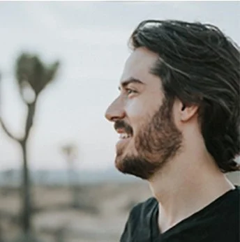 Side profile of a man with dark hair and beard smiling outdoors with trees in the background.