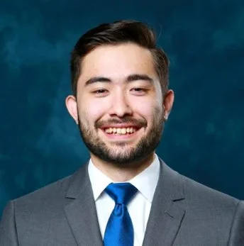 A young man with dark hair and a beard wearing a gray suit, white shirt, and blue tie, smiling against a dark blue background.