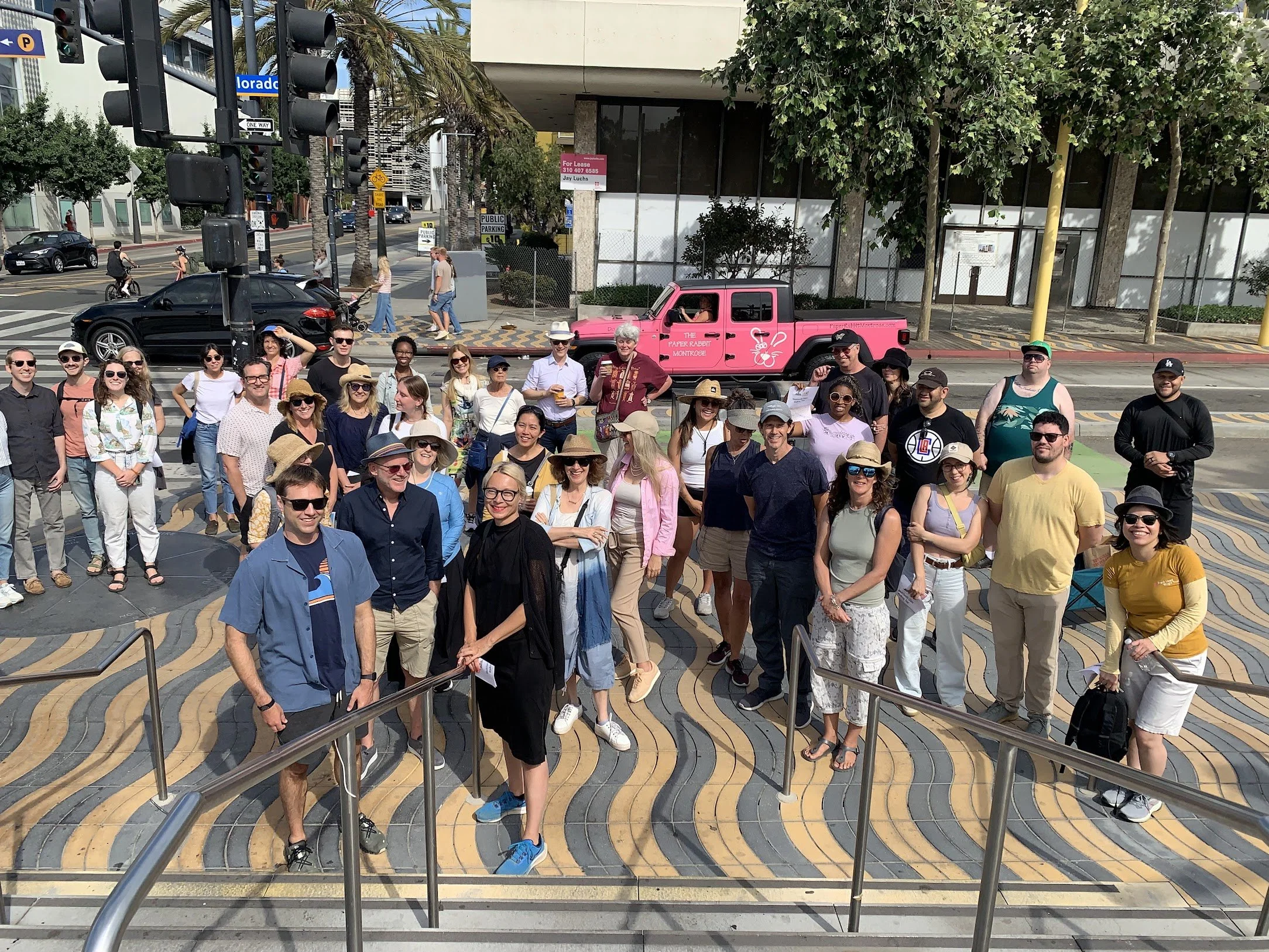 Group of people standing on a colorful, striped patterned sidewalk with a pink vehicle behind them. People are smiling and wearing casual clothing, some are wearing hats and sunglasses. The background shows buildings, trees, and street signs.
