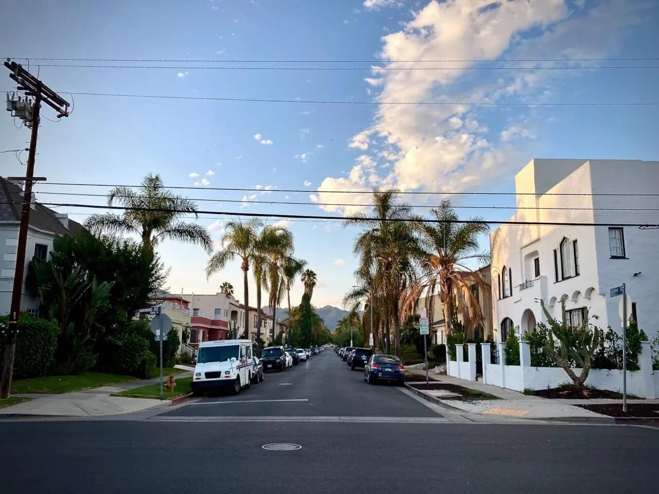 Residential street lined with palm trees, white and pastel-colored houses, parked cars along the sides, and a mountain view in the distance under a partly cloudy sky.