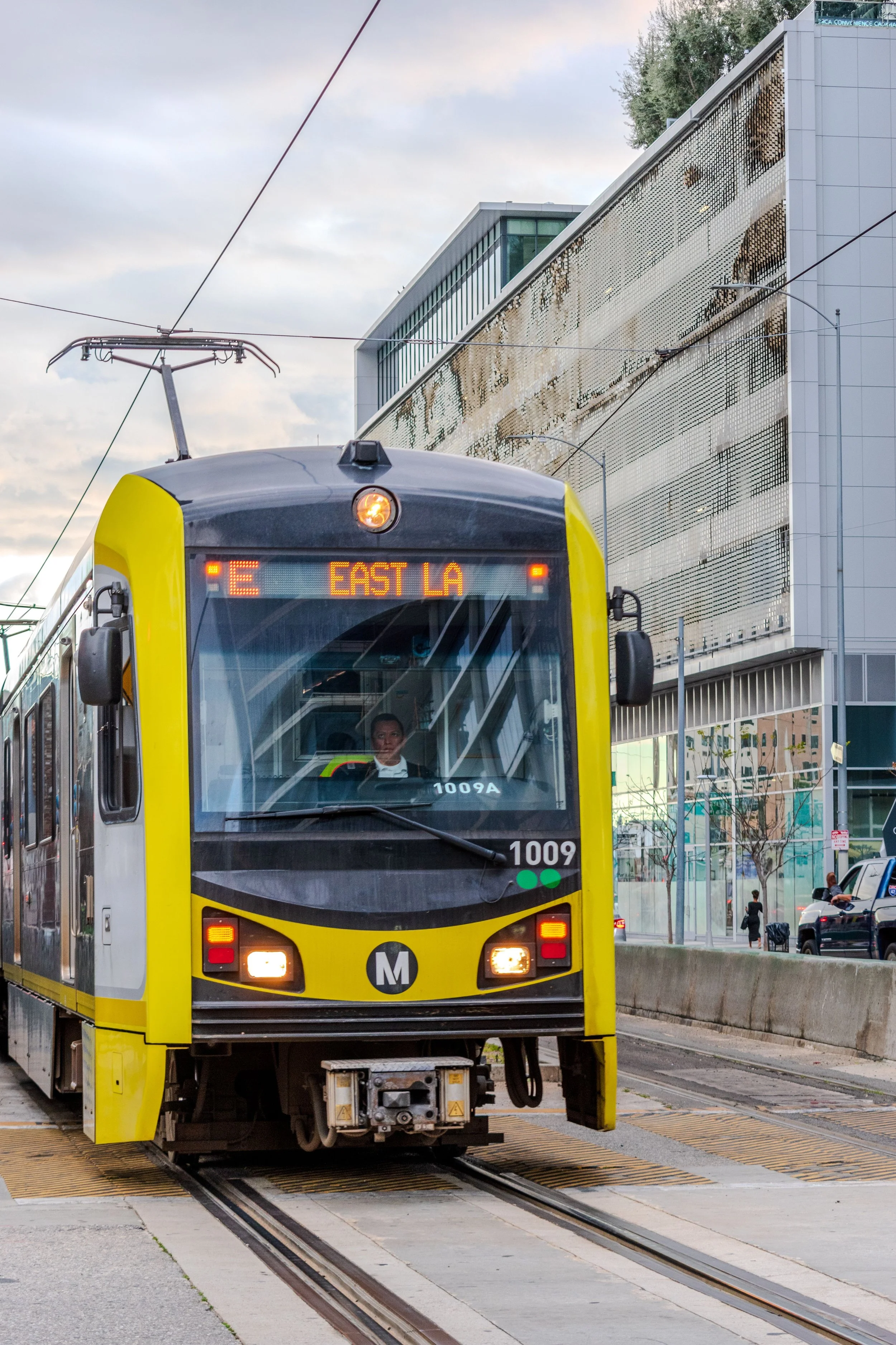 A yellow light rail train with digital display showing route information, approaching a station with modern glass buildings and overhead electric wires, under a cloudy sky.