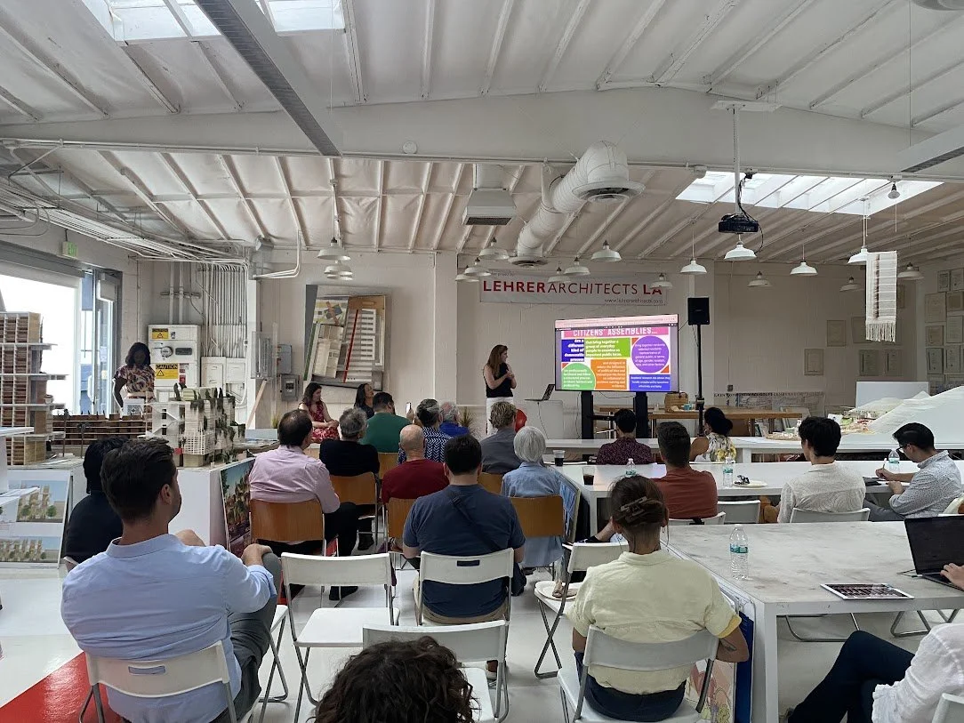 A speaker and a woman standing in front of a seated audience in an industrial-style room with white walls, exposed ductwork, and a large digital screen displaying a presentation on citizens' assemblies.