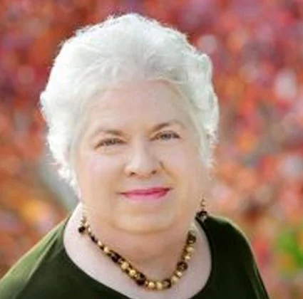 A woman with short white hair, wearing a black top and a gold beaded necklace, smiling outdoors with fall foliage in the background.