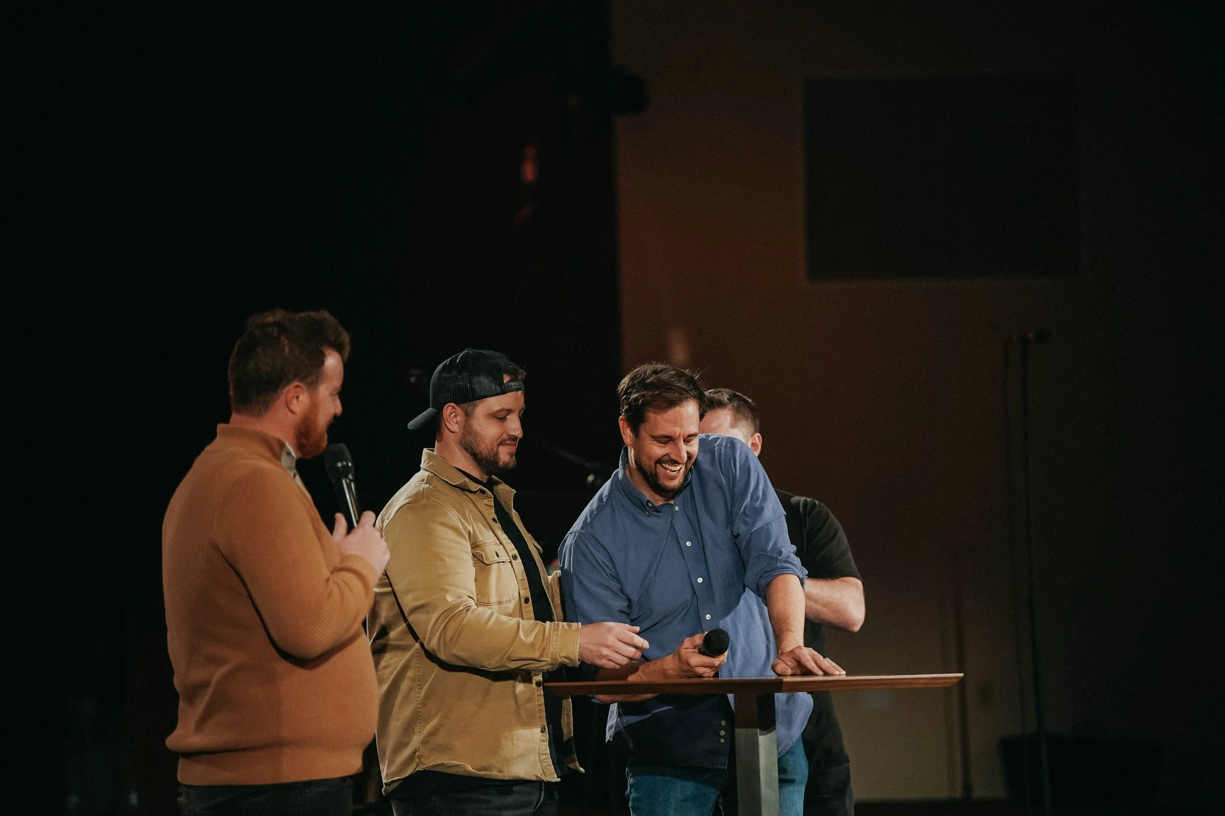 Four men are standing on a stage, engaged in conversation or activity, with dark background and a wooden table or podium in front of them. One man on the left is holding a microphone, and the man on the right is smiling while looking down, possibly at something on the table.