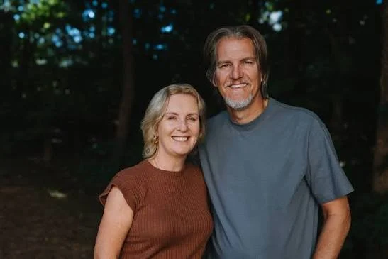 A smiling middle-aged woman with blonde hair and a man with gray hair and beard, both standing outdoors at dusk or evening.