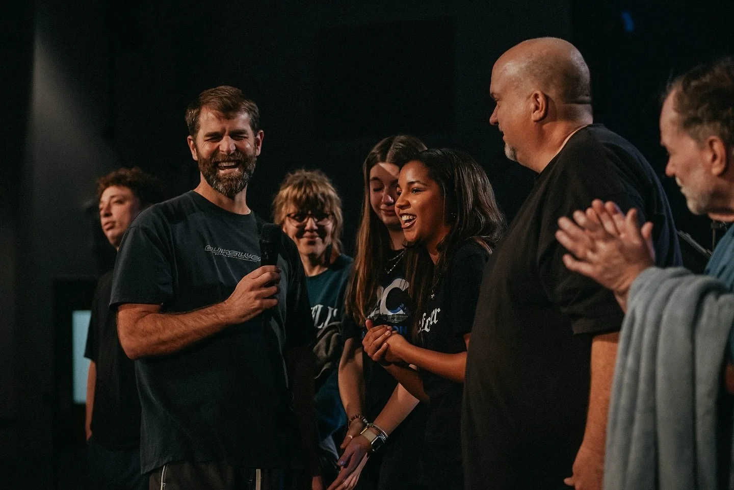 Group of people on stage, with one man holding a microphone, smiling, and engaging with women who are smiling and clapping.