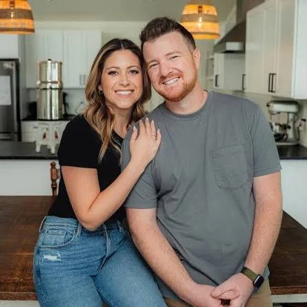 A smiling couple sitting together in a kitchen, with the woman resting her hand on the man's shoulder.