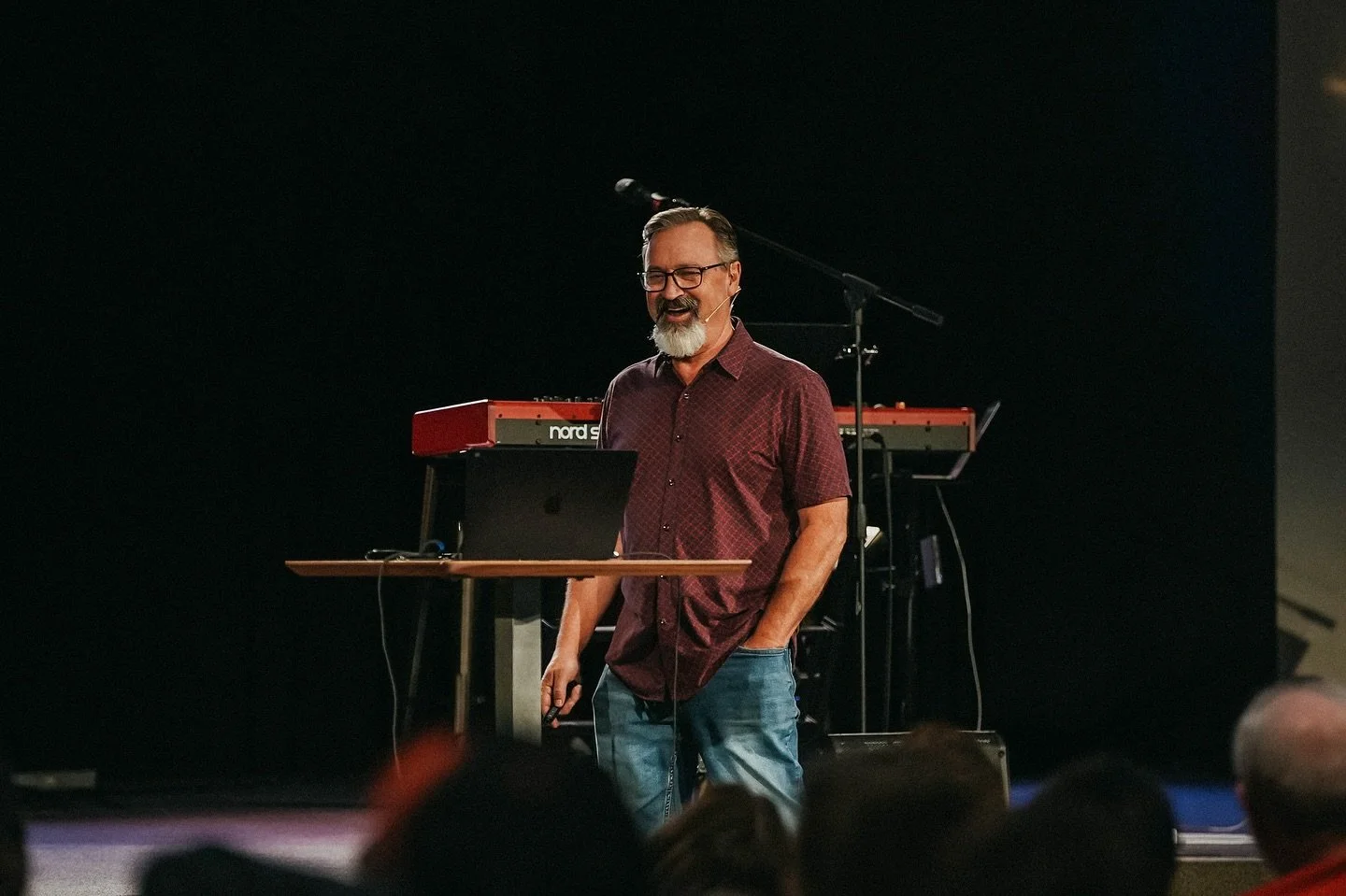 Man with glasses and a beard speaking on stage with musical equipment behind him.