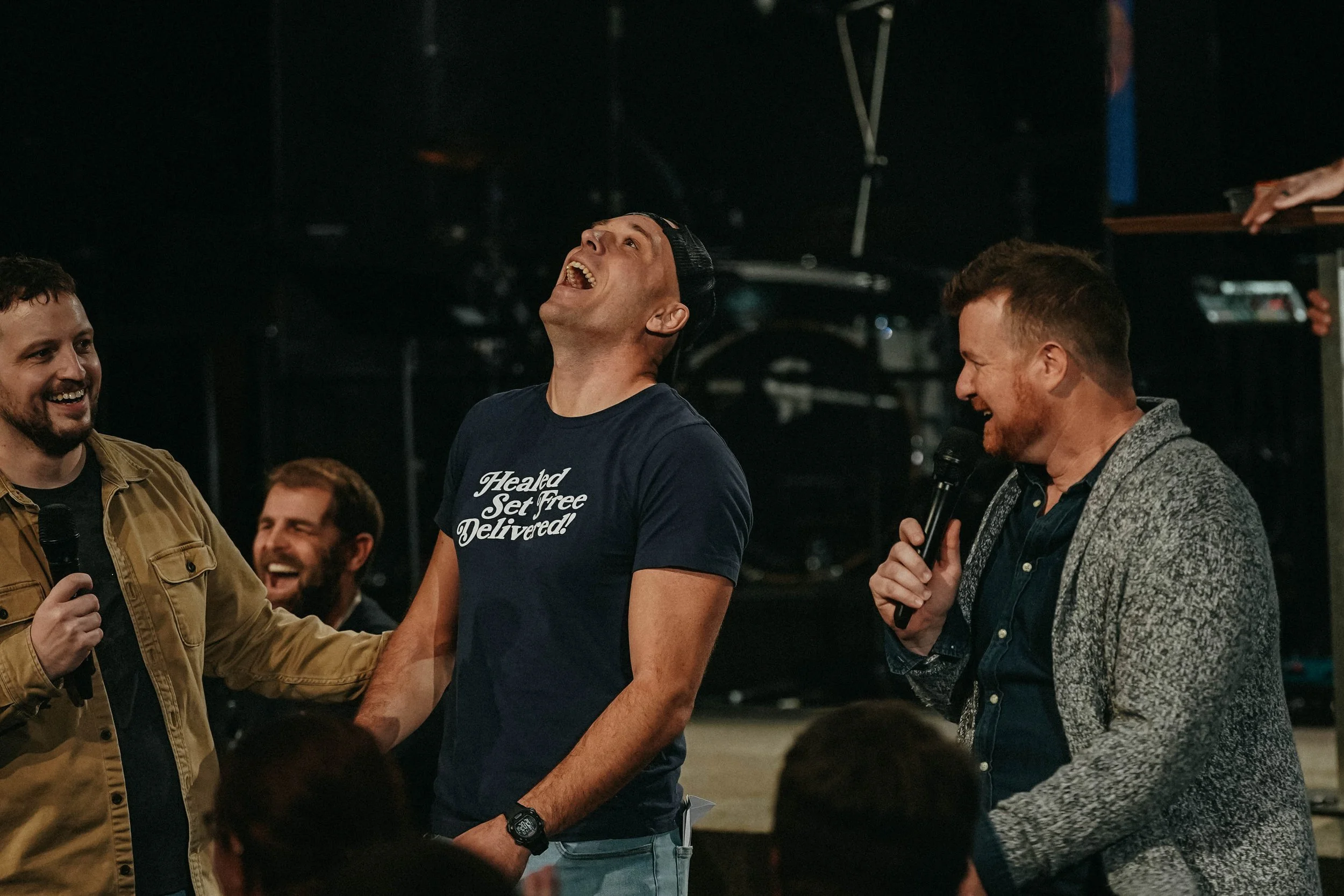 Four men are laughing and talking in an indoor setting, with three of them holding microphones. The man in the middle is wearing a navy T-shirt with white text that reads, 'Healed Set Free Delivered!'. The man on the right is wearing a gray blazer and speaking into a microphone. The man on the left is wearing a tan jacket and smiling. There are blurred people and equipment in the background.