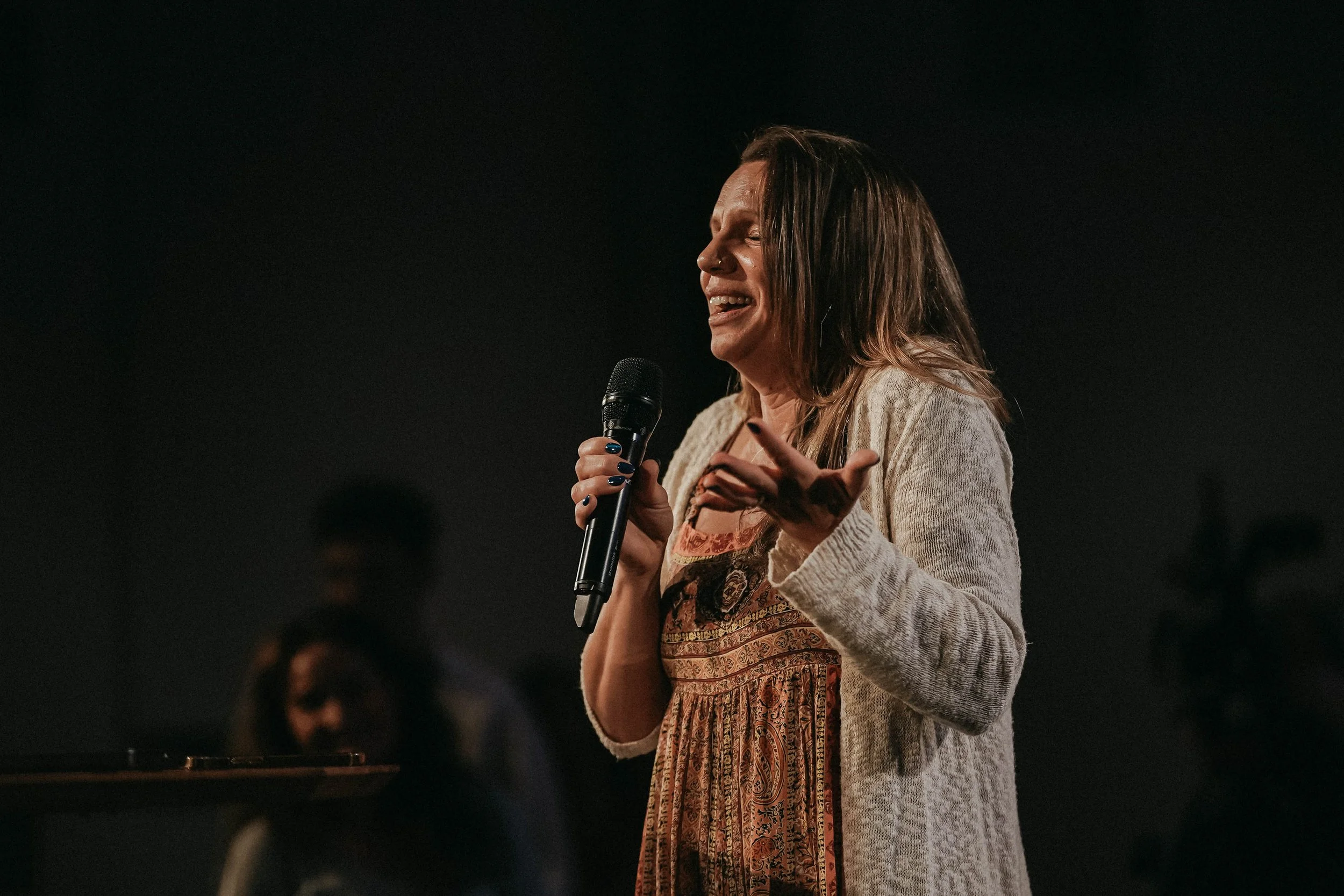 A woman singing or speaking into a microphone on stage with a dark background.