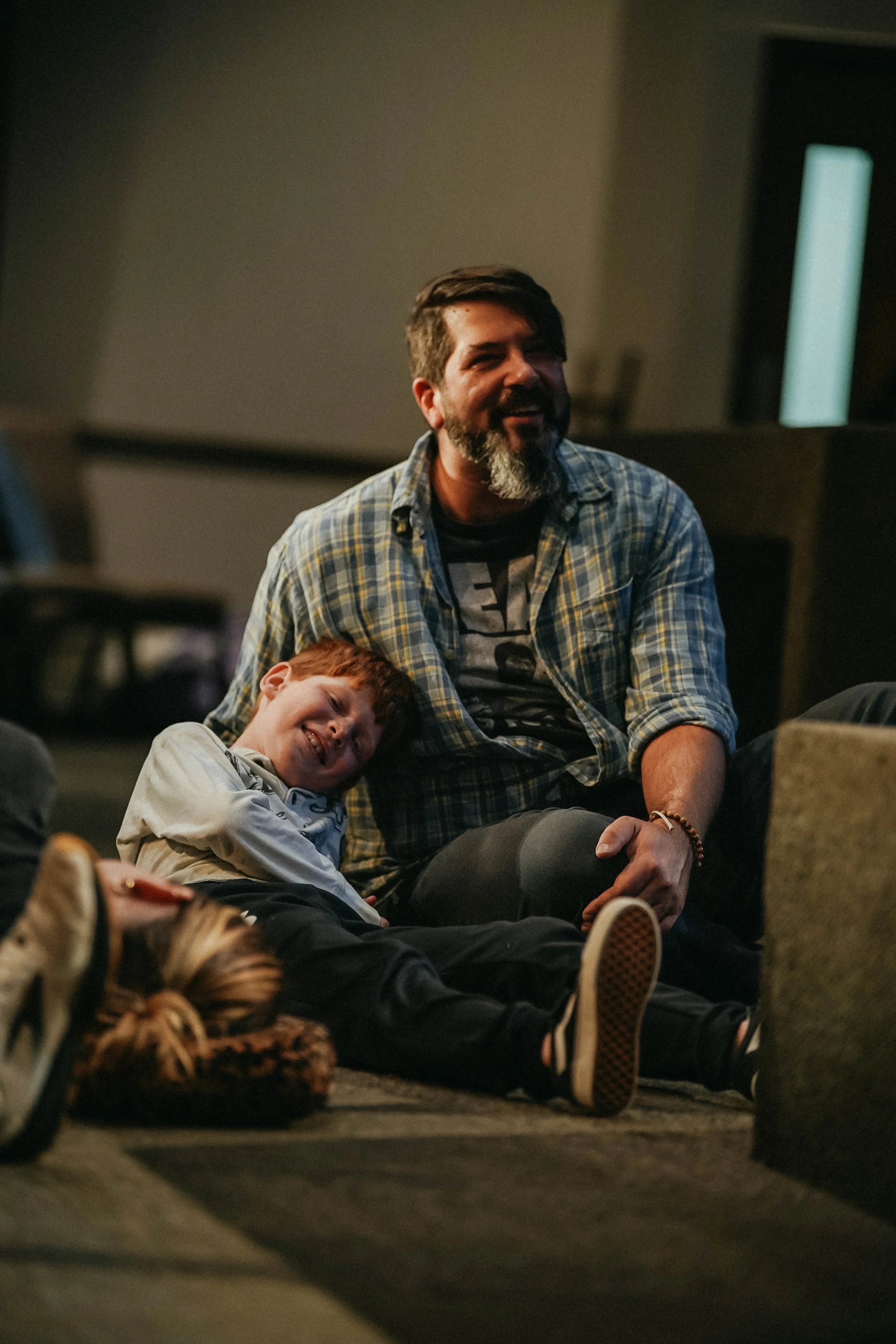 A man with dark hair, a beard, and a mustache, wearing a plaid shirt, is sitting on the floor, smiling, with a young boy with red hair and a light-colored jacket leaning against him, also smiling and relaxed.