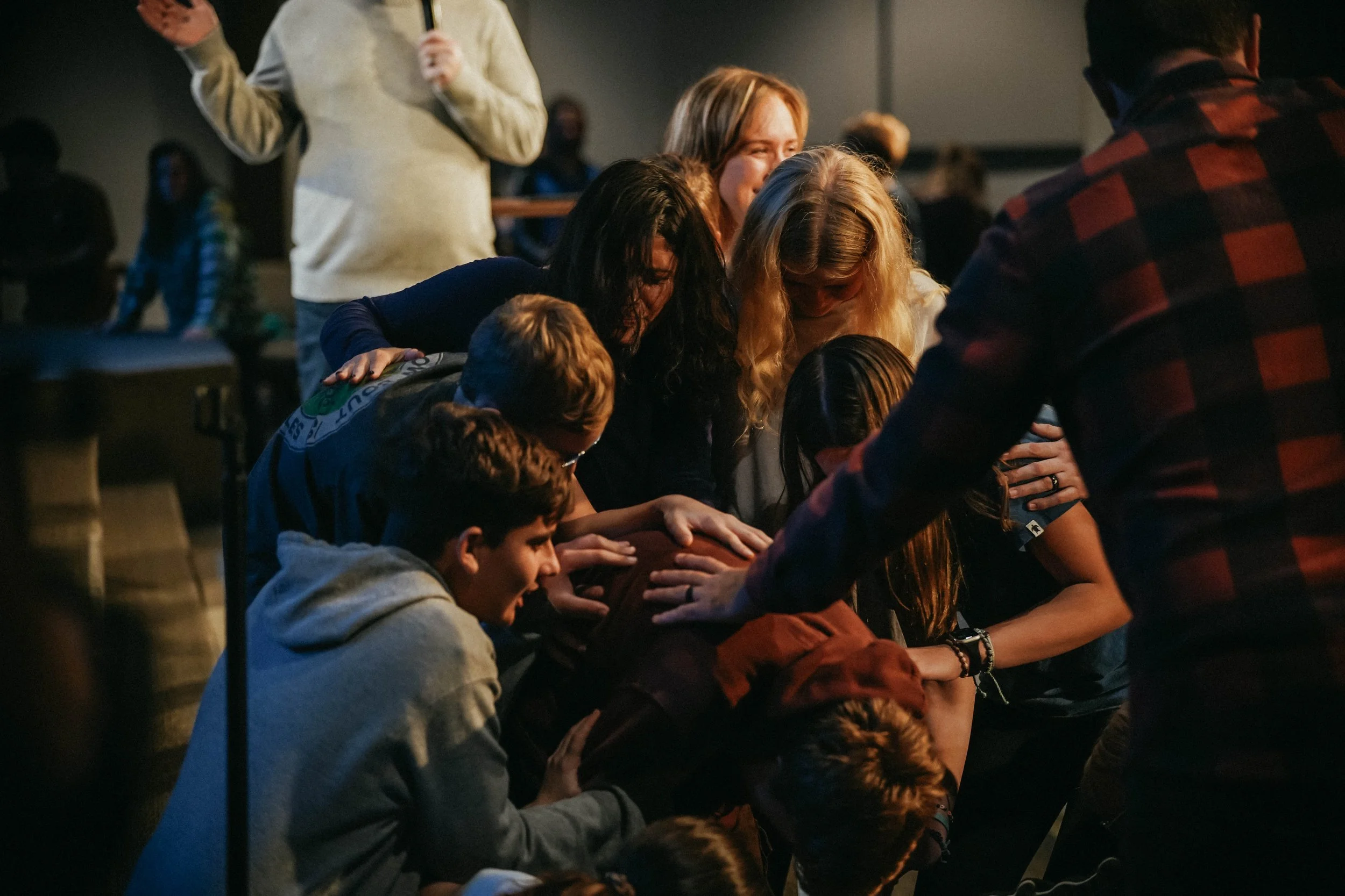 People kneeling with heads bowed and hands placed on each other's shoulders, creating a prayer circle in a dimly lit room, with others standing and observing in the background.