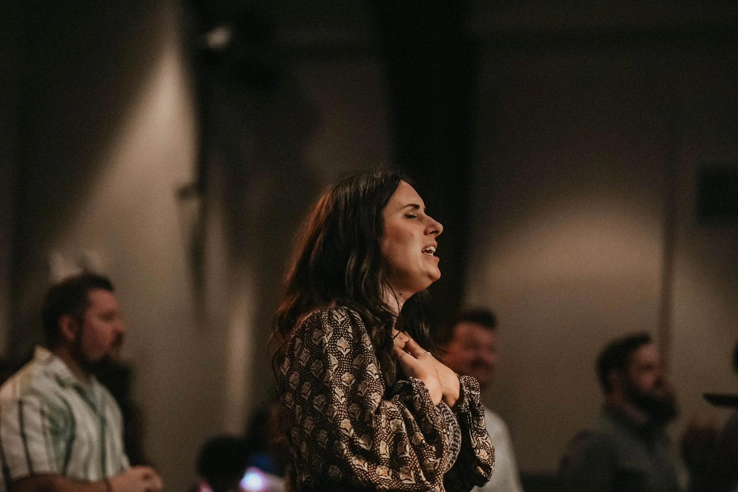 A woman with long dark hair and a patterned dress appears to be singing with her eyes closed at an event, surrounded by people in the background.