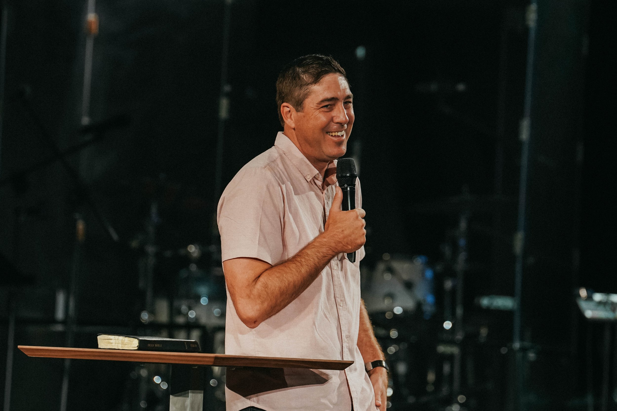 A man smiling and speaking into a microphone during a presentation or speech, with a Bible and a table in front of him.