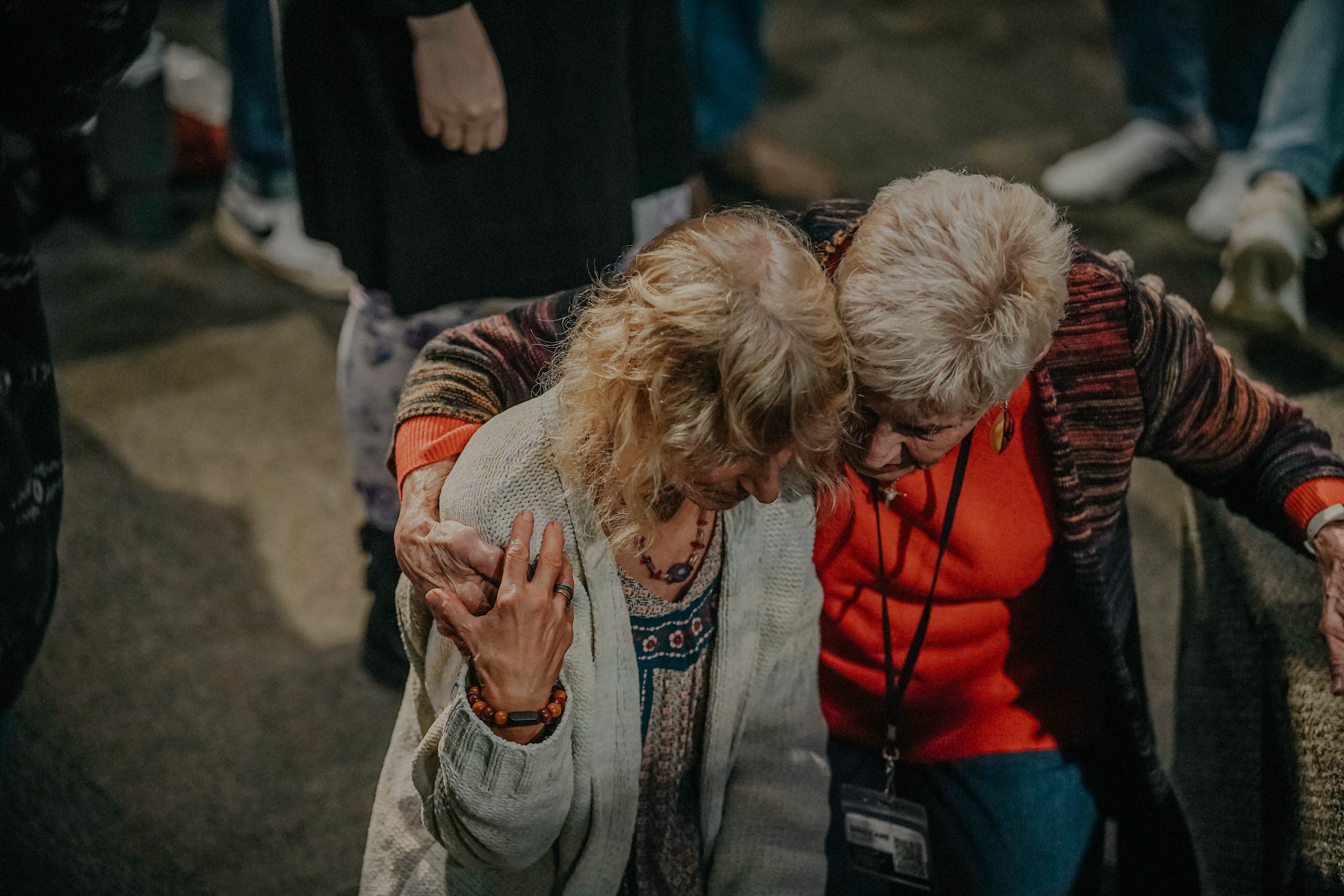 Two elderly women embracing in a hug at an indoor event, surrounded by other attendees.
