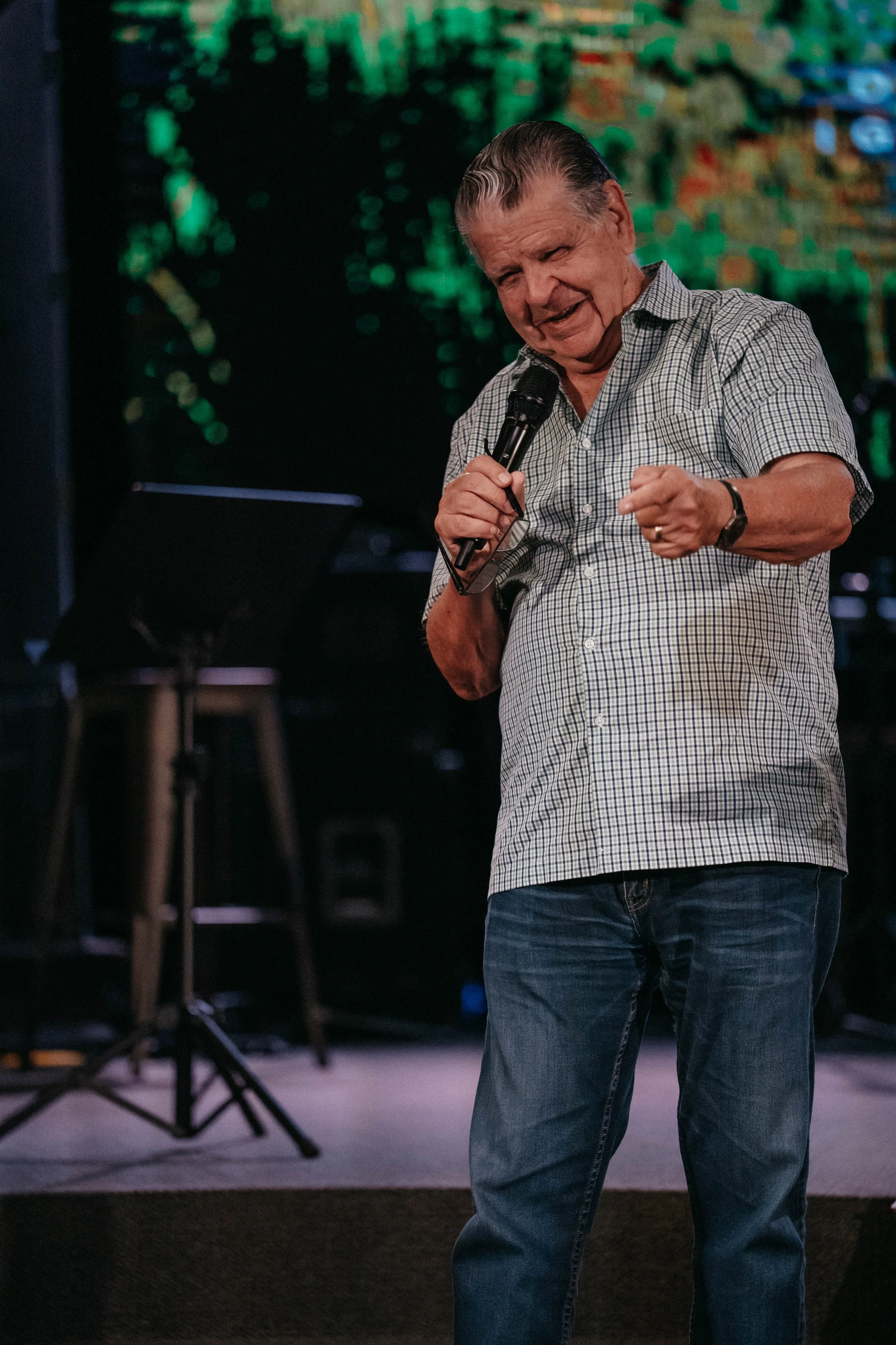 An elderly man holding a microphone, speaking or singing, with a colorful digital background and a music stand behind him.