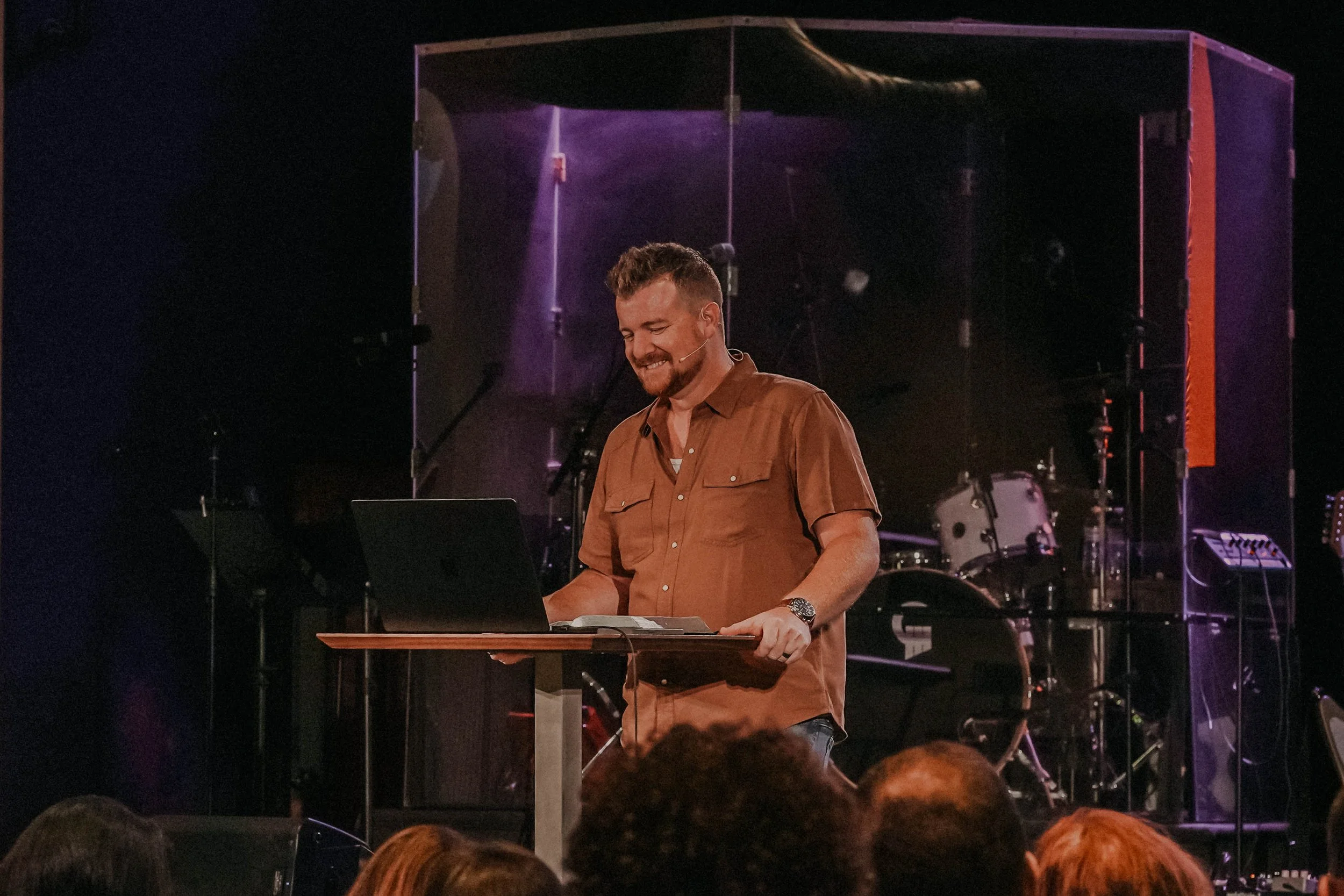 A man smiling while playing a keyboard on stage with musical instruments behind him.
