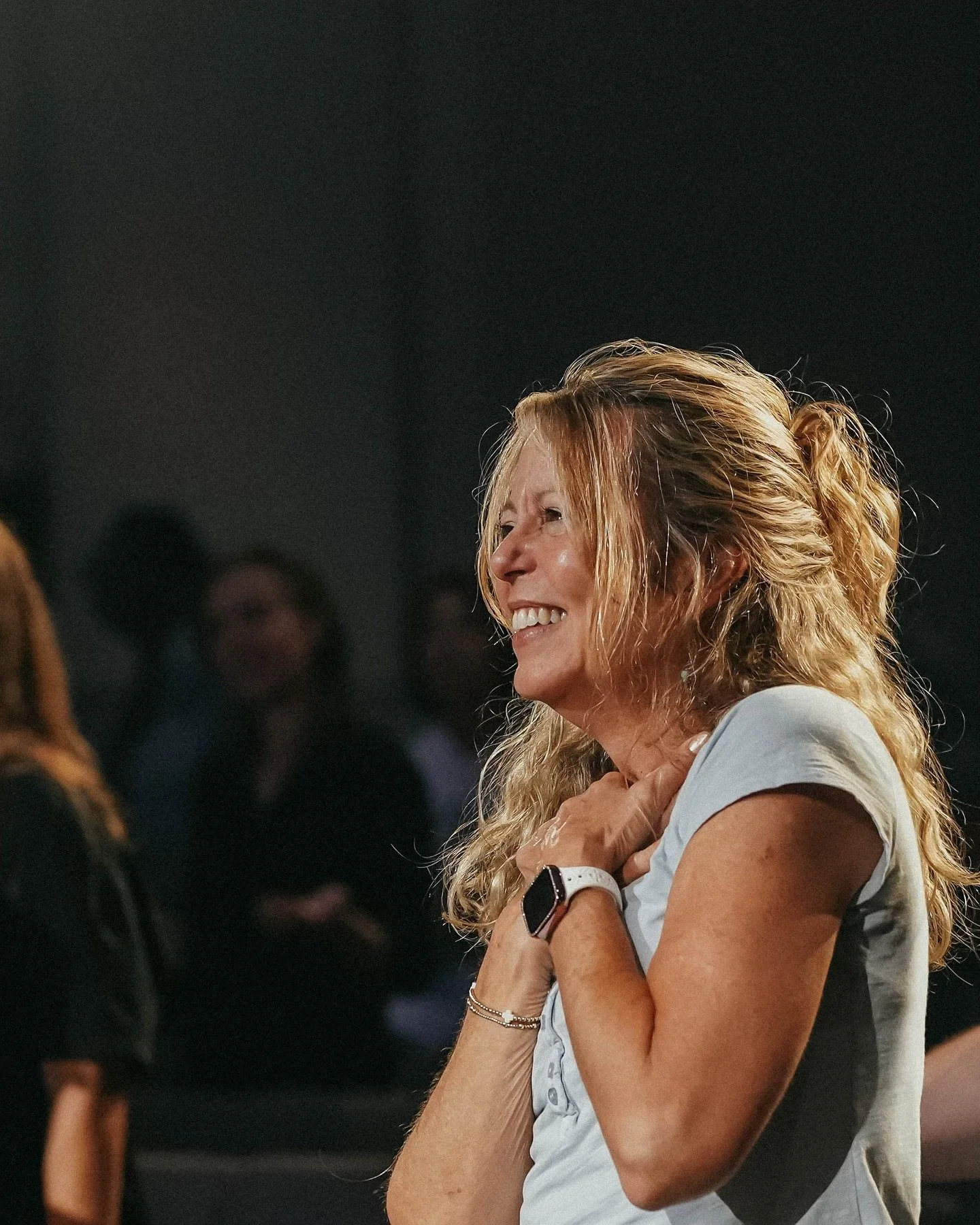 A woman with long, wavy blonde hair smiling and holding her hand to her chest in a lively indoor setting with blurred people in the background.
