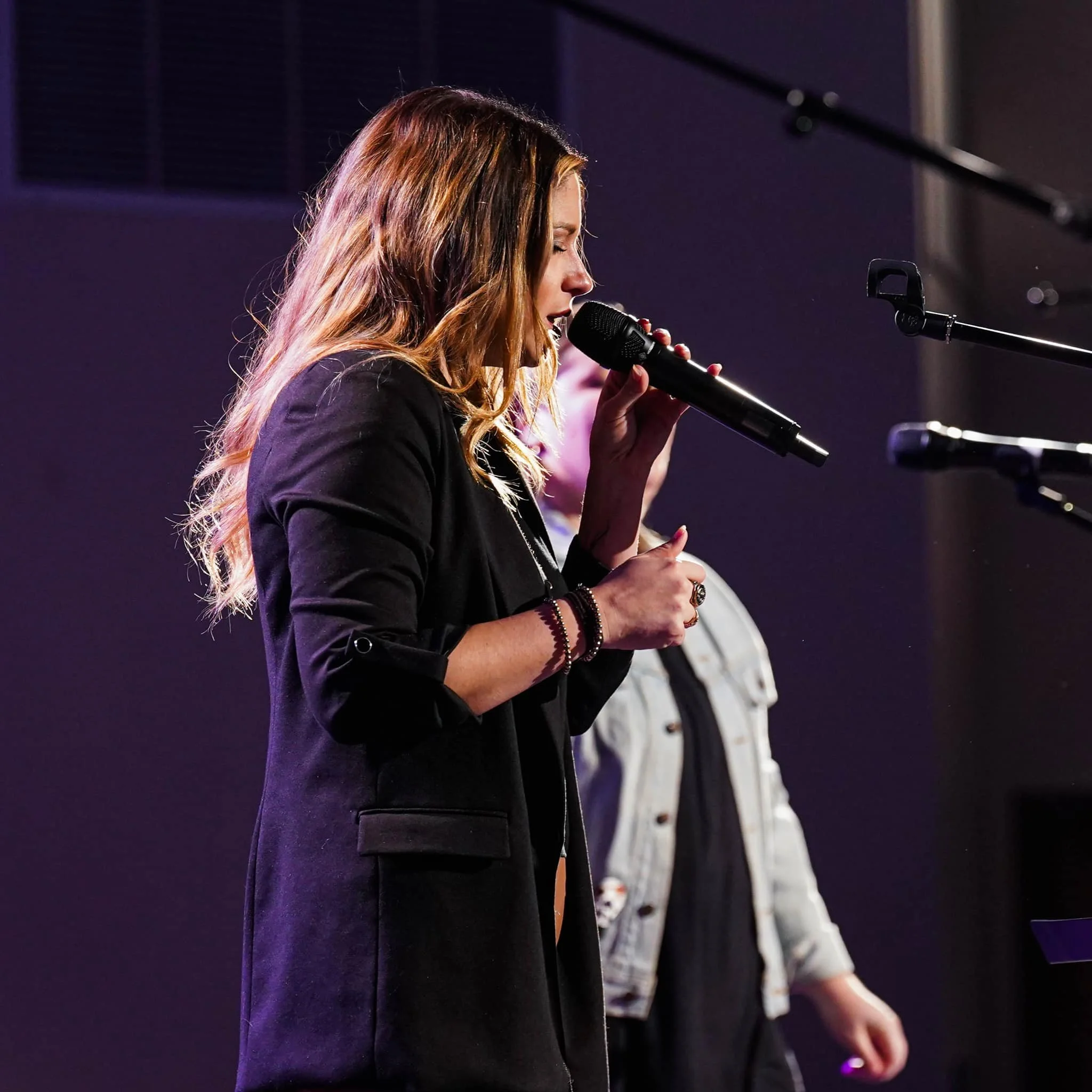 A woman with long, wavy, reddish-brown hair singing into a microphone on stage, wearing a black blazer, with stage lights illuminating her hair and face, and a dark background