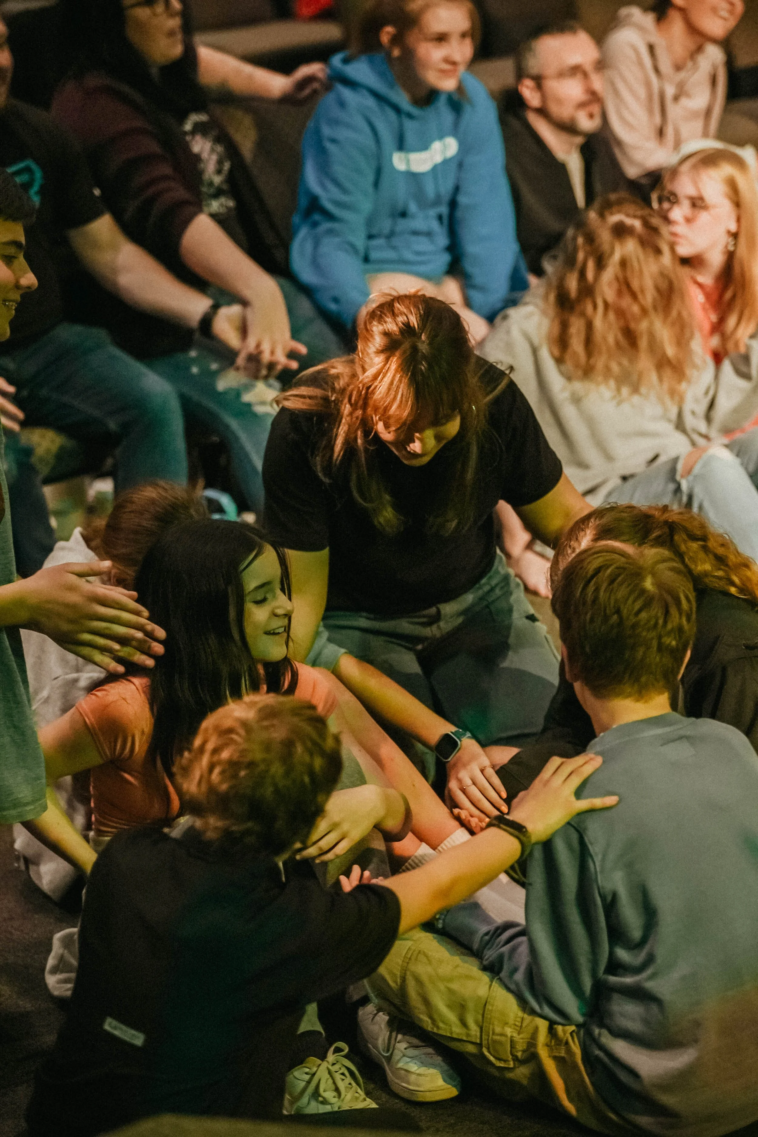 Group of young people sitting in a circle, engaging in a shared activity, with some reaching towards the center, in a casual indoor setting.