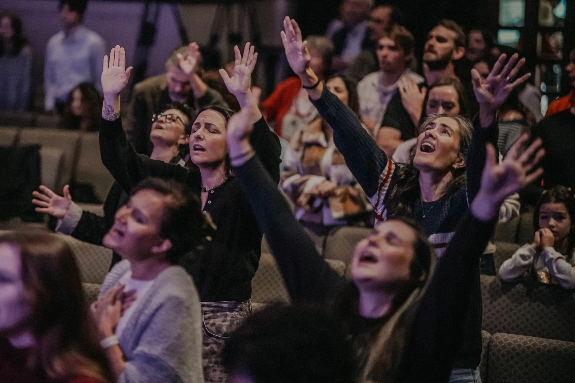 A diverse group of people, including women and children, gathered in a darkened auditorium with some raising their hands and looking upward, possibly during a religious or spiritual event.