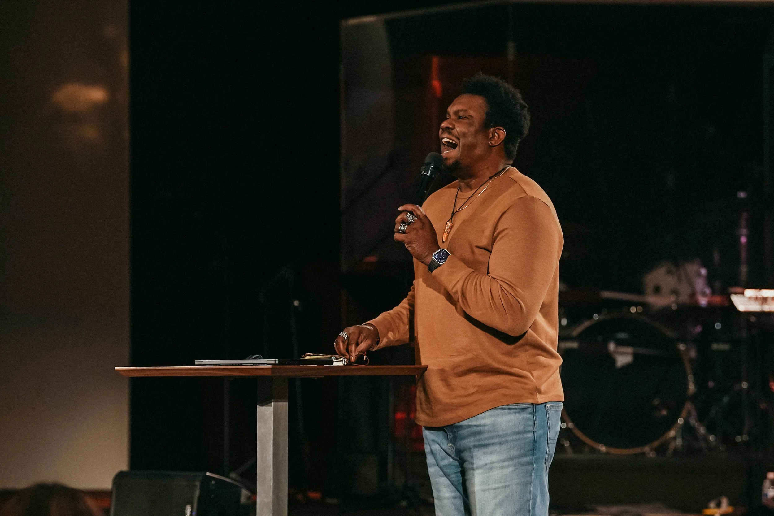 A man with dark curly hair singing into a microphone at a performance, wearing a tan long-sleeve shirt and jeans, standing at a table with a laptop in a dimly lit venue.