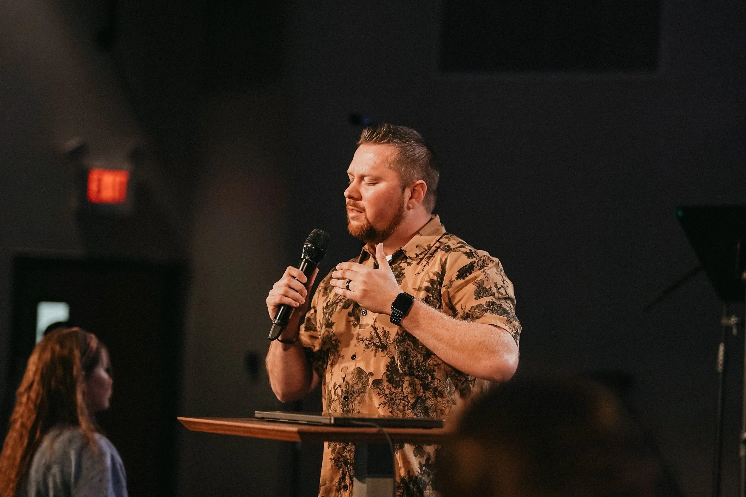 A man with short hair and a beard is singing into a microphone with his eyes closed, wearing a tan, patterned shirt, while standing at a podium during a presentation or speech in a dimly lit room.