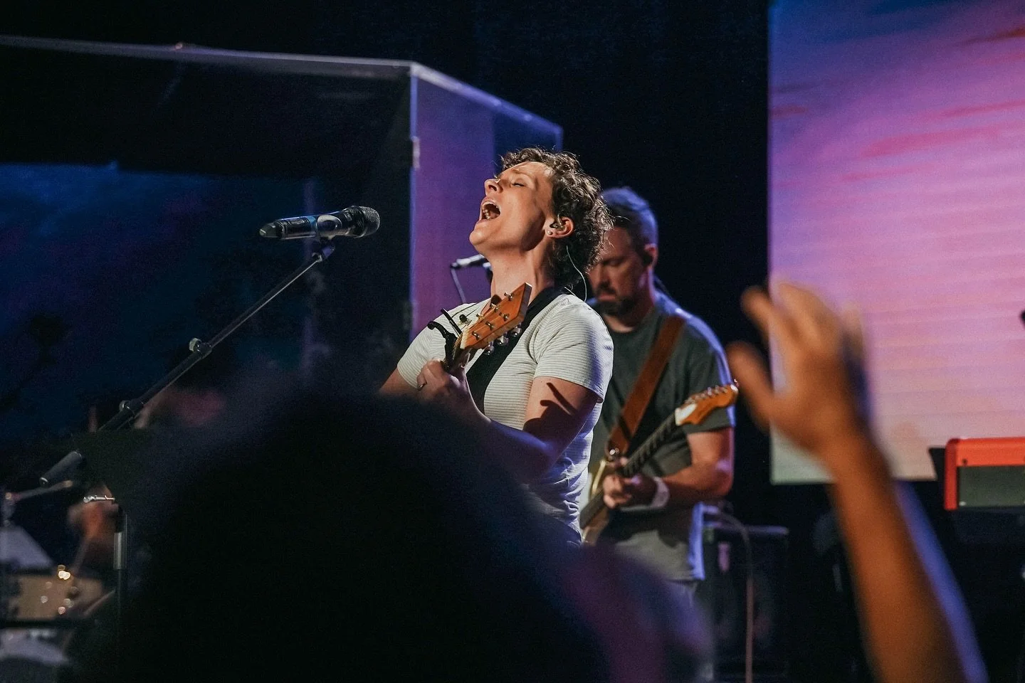 A woman singing passionately while playing an acoustic guitar on stage, with a man playing an electric guitar in the background, in a dimly lit concert setting with people raising their hands.
