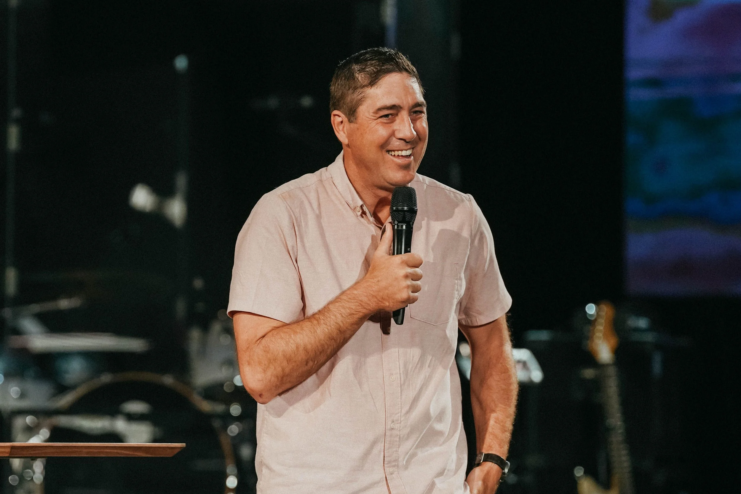 Man smiling and speaking into a microphone during a presentation or speech, wearing a light-colored short-sleeve shirt, with musical instruments and equipment in the background.