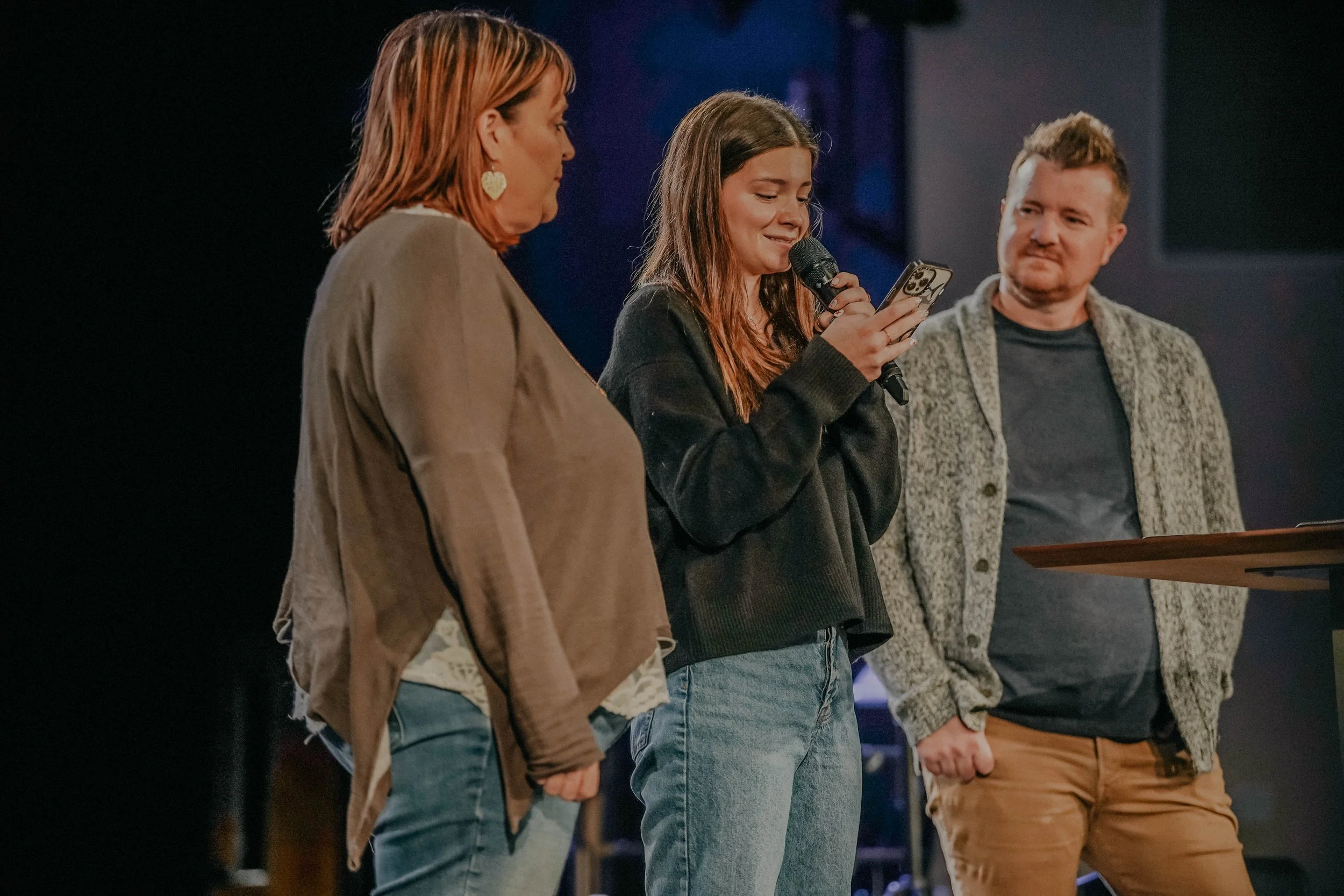 Three people standing on a stage, with the woman in the middle looking at her phone and speaking into a microphone, while the other two look on.