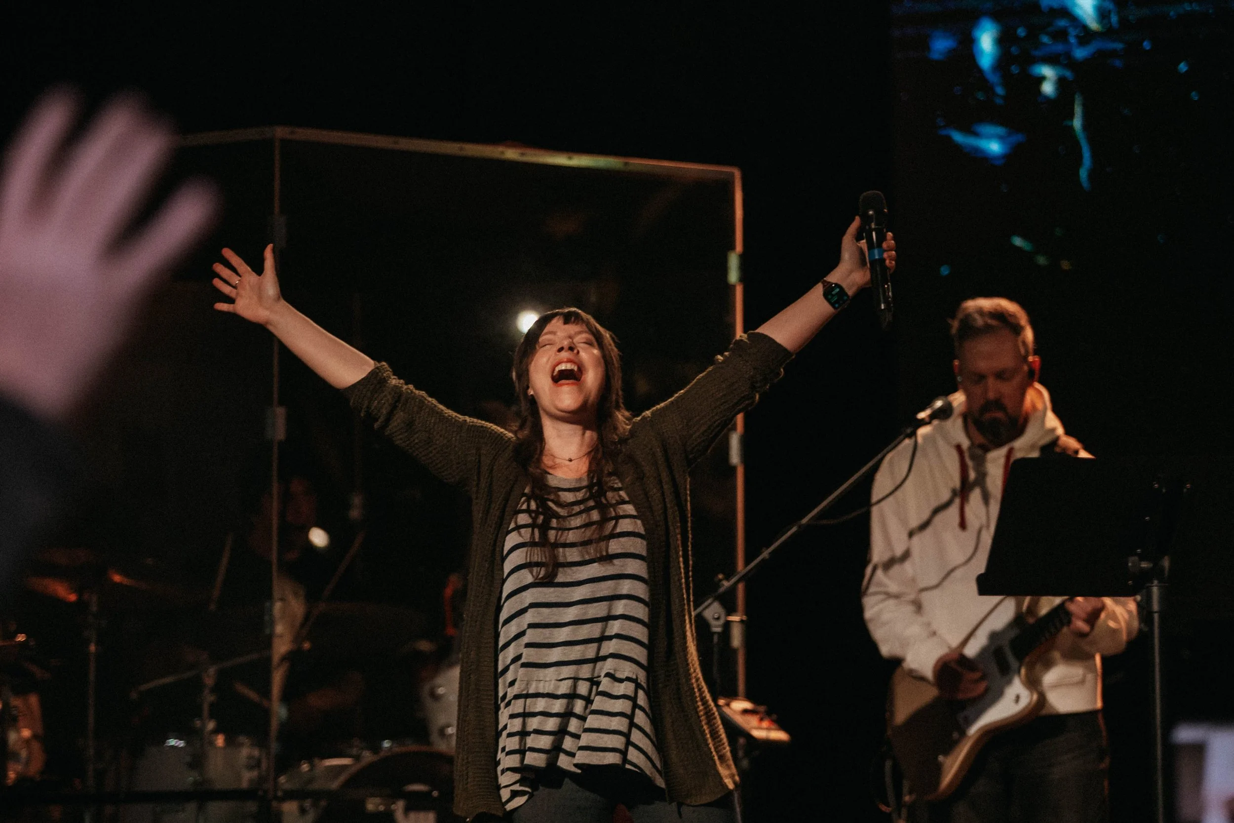 A young woman with brown hair and striped shirt singing with arms outstretched, holding a microphone, on stage with band members playing guitar and keyboard in the background, at night under stage lights.