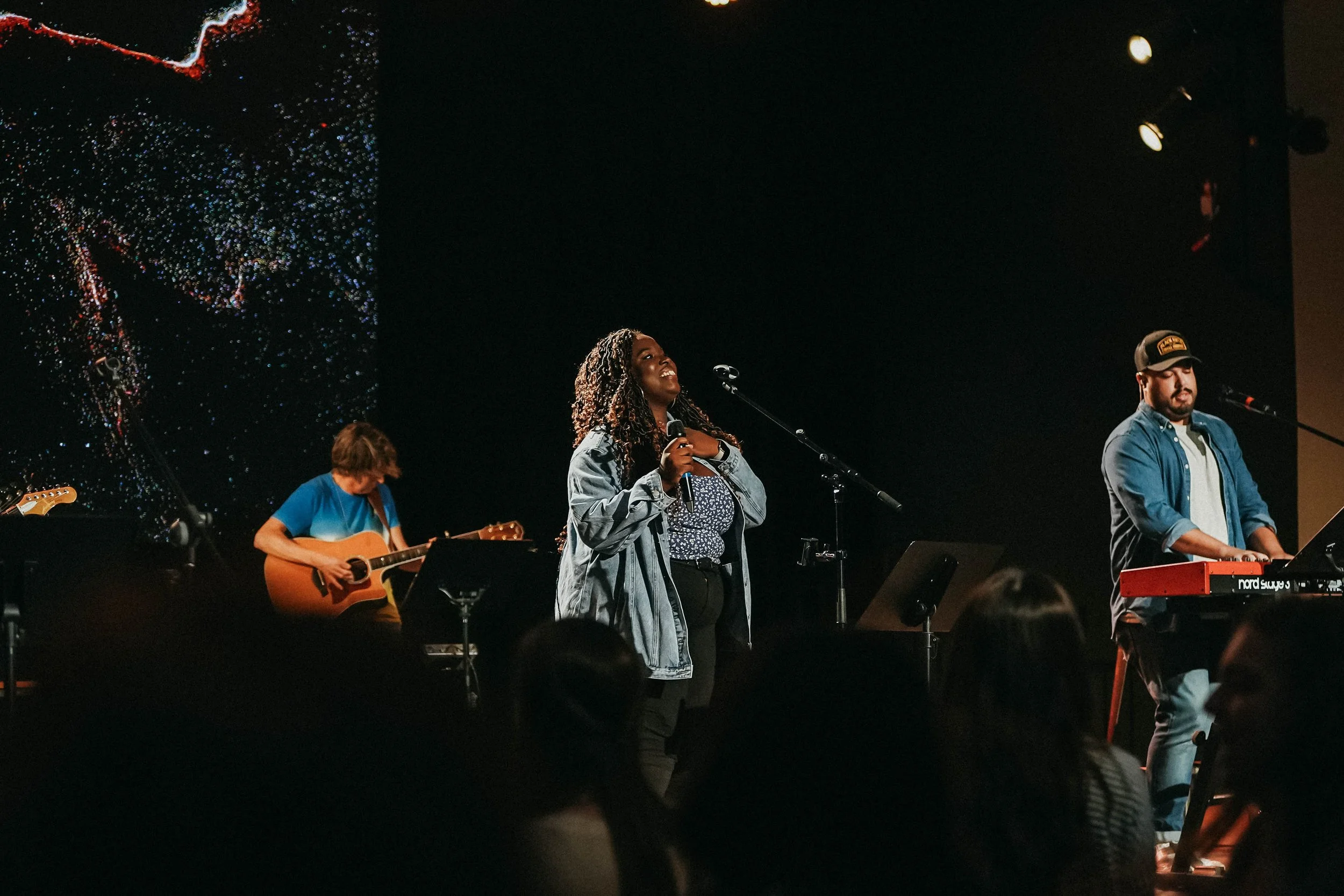 A group of musicians performing on stage with a female vocalist holding a microphone. The band includes a guitarist and a keyboardist, with stage lights and a black backdrop.