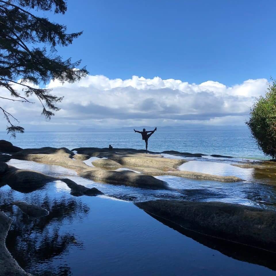Person standing on rocks near the water with arms outstretched in a yoga pose, overlooking a lake and cloudy sky during daytime.