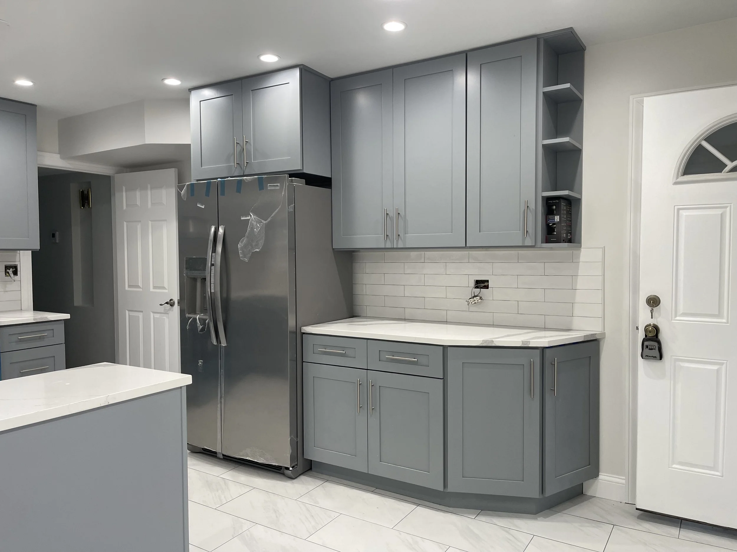A modern kitchen with blue cabinets, white countertops, and a stainless steel refrigerator. Recessed ceiling lights and a white tiled backsplash are visible. A microwave is placed in a corner shelf in the cabinet unit to the right of the fridge.