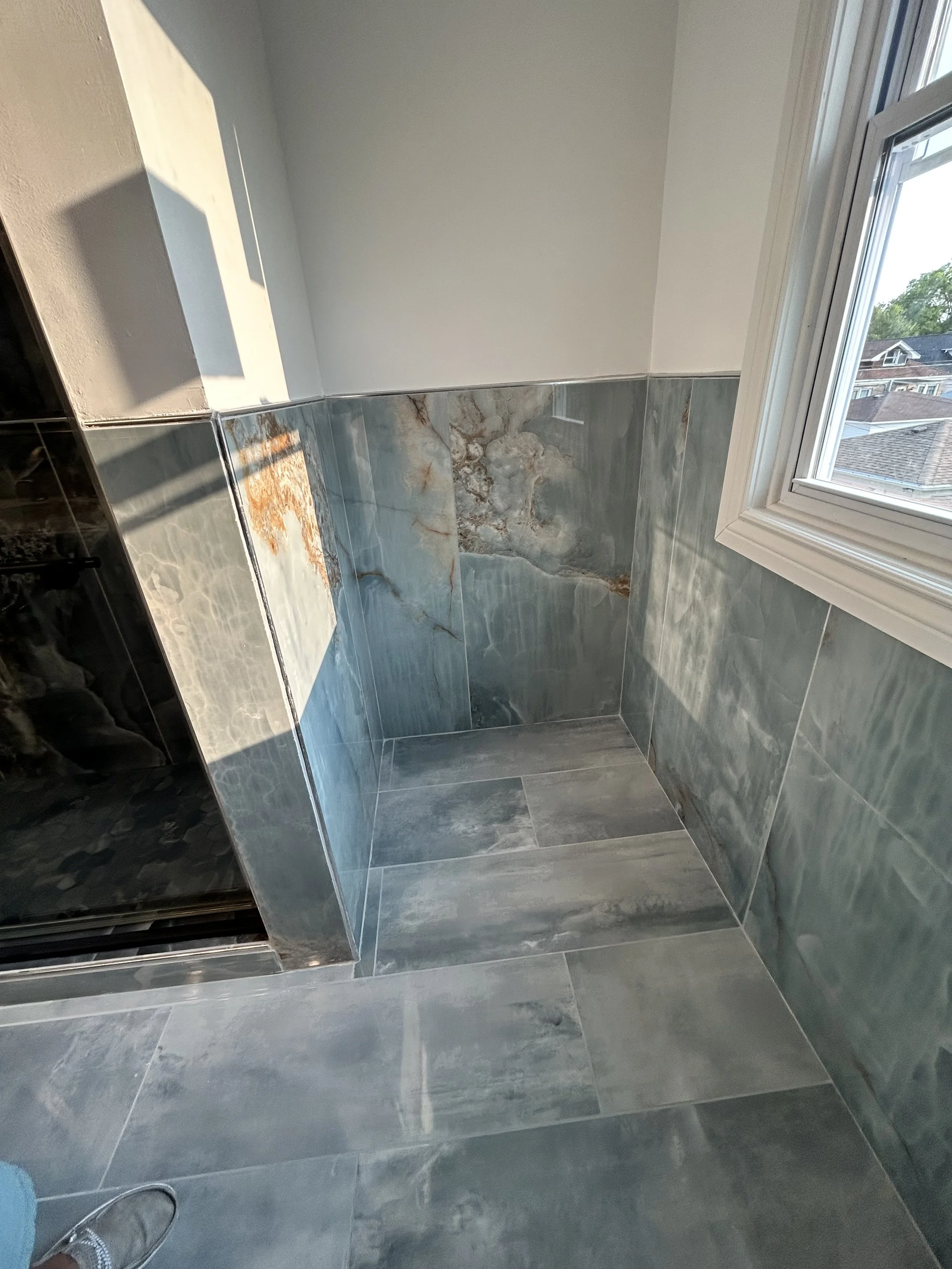 Interior of a room with gray marble tiles on the lower half of the walls and floor, a window with white trim on the right wall, and a beige upper wall. The photo is taken from someone standing inside the room, with part of their shoe visible in the l