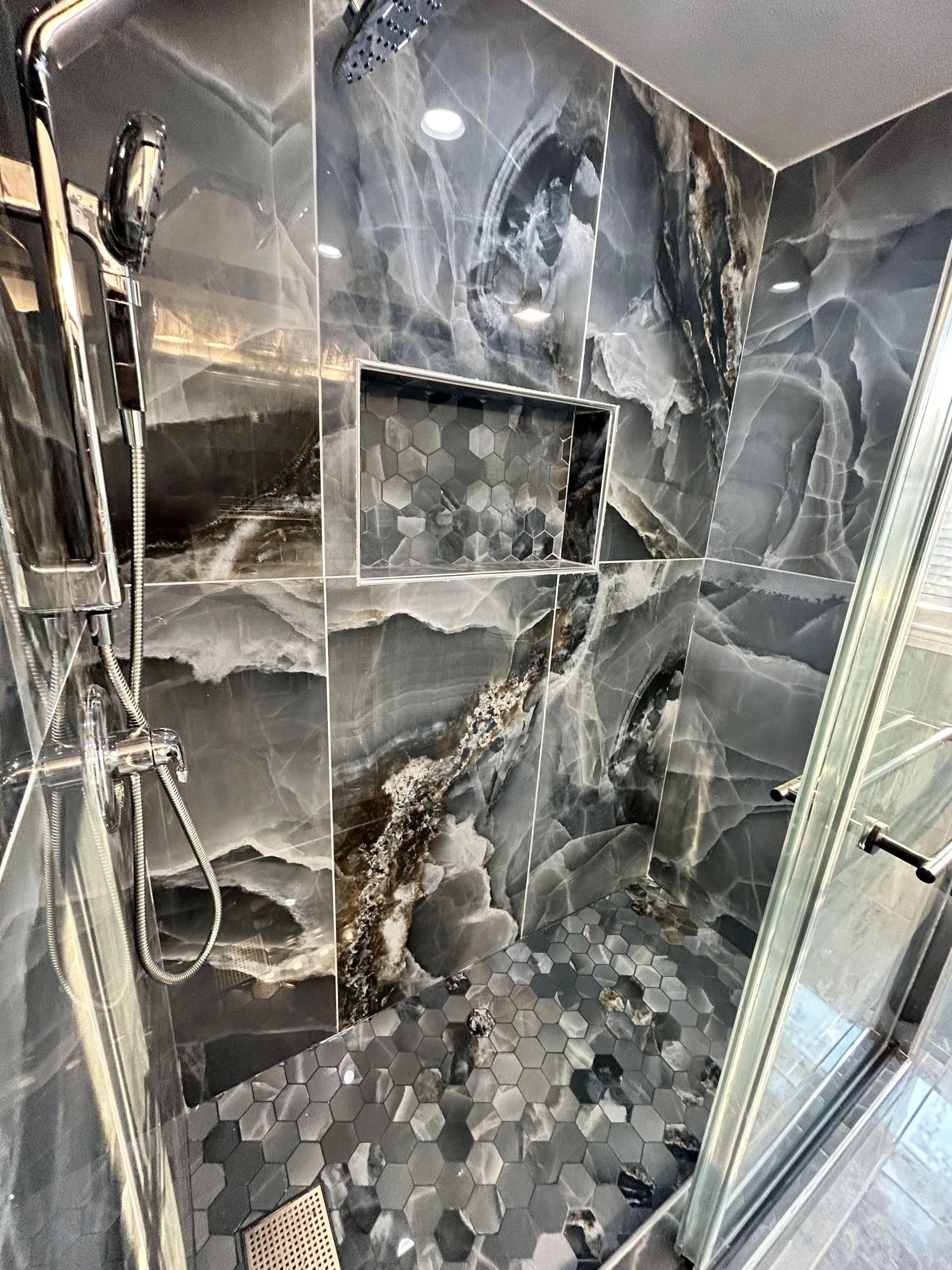 Marble tile shower with hexagon floor tiles, a built-in niche with matching marble and small square tiles, and a rainfall showerhead, glass door and stainless steel fixtures.