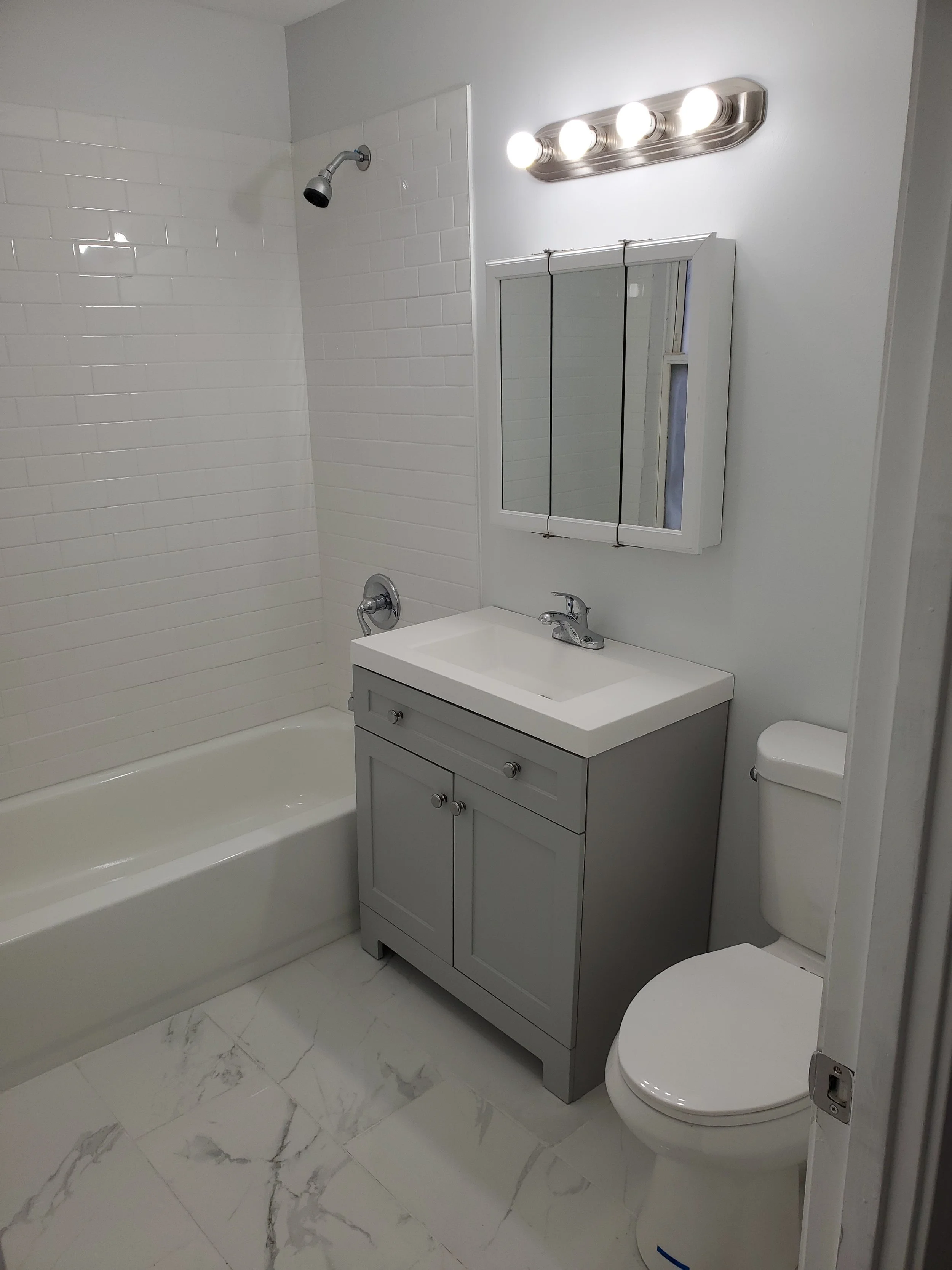 A small bathroom featuring a bathtub with white tiled walls, a gray vanity with a white sink, a mirrored medicine cabinet, a toilet, and marble-patterned floor tiles. There is a bright vanity light fixture above the mirror.