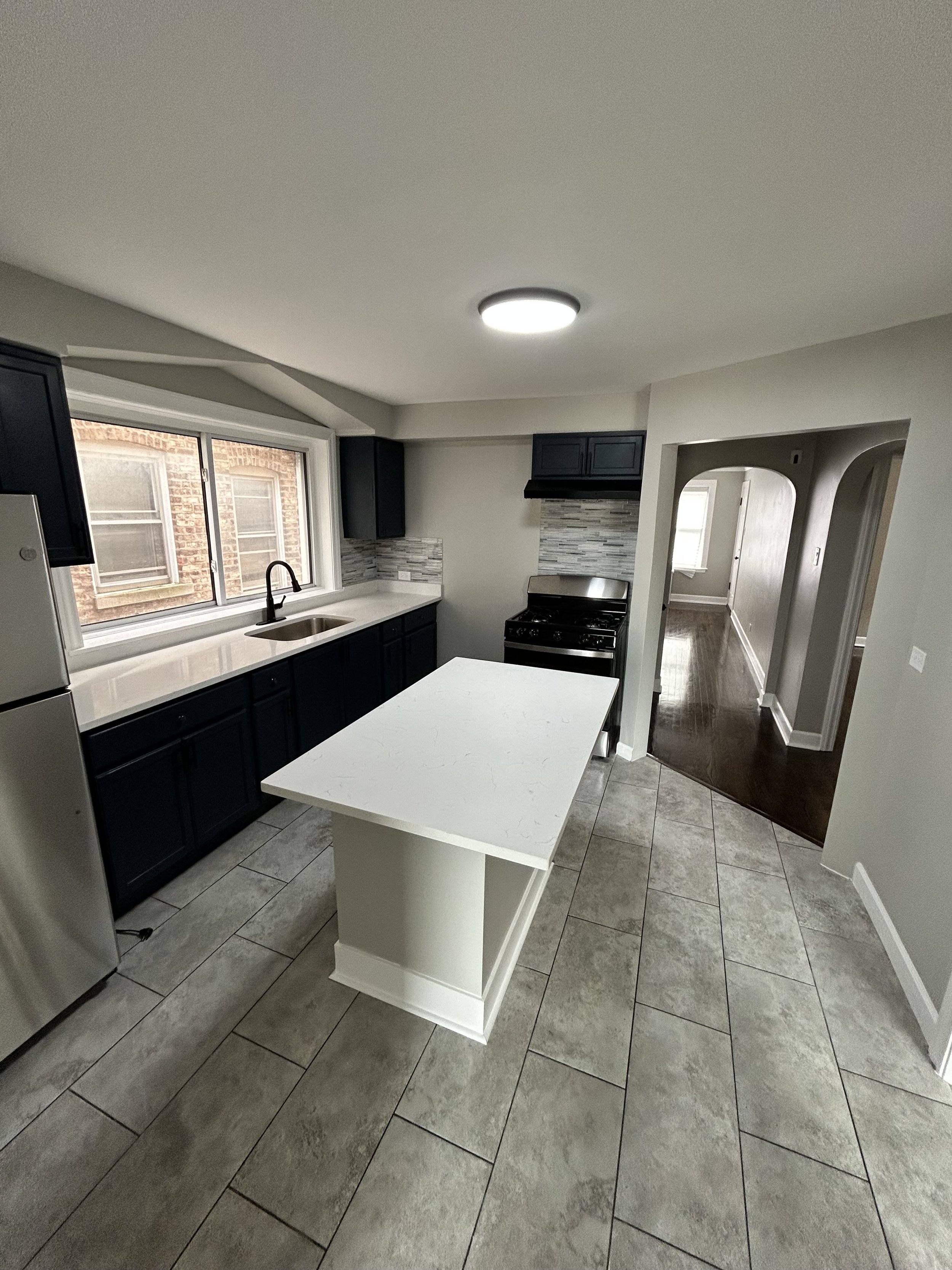 Kitchen with white island, dark blue cabinets, gray and white backsplash, window above sink, stainless steel stove, tiled floor, and passageway to other rooms.
